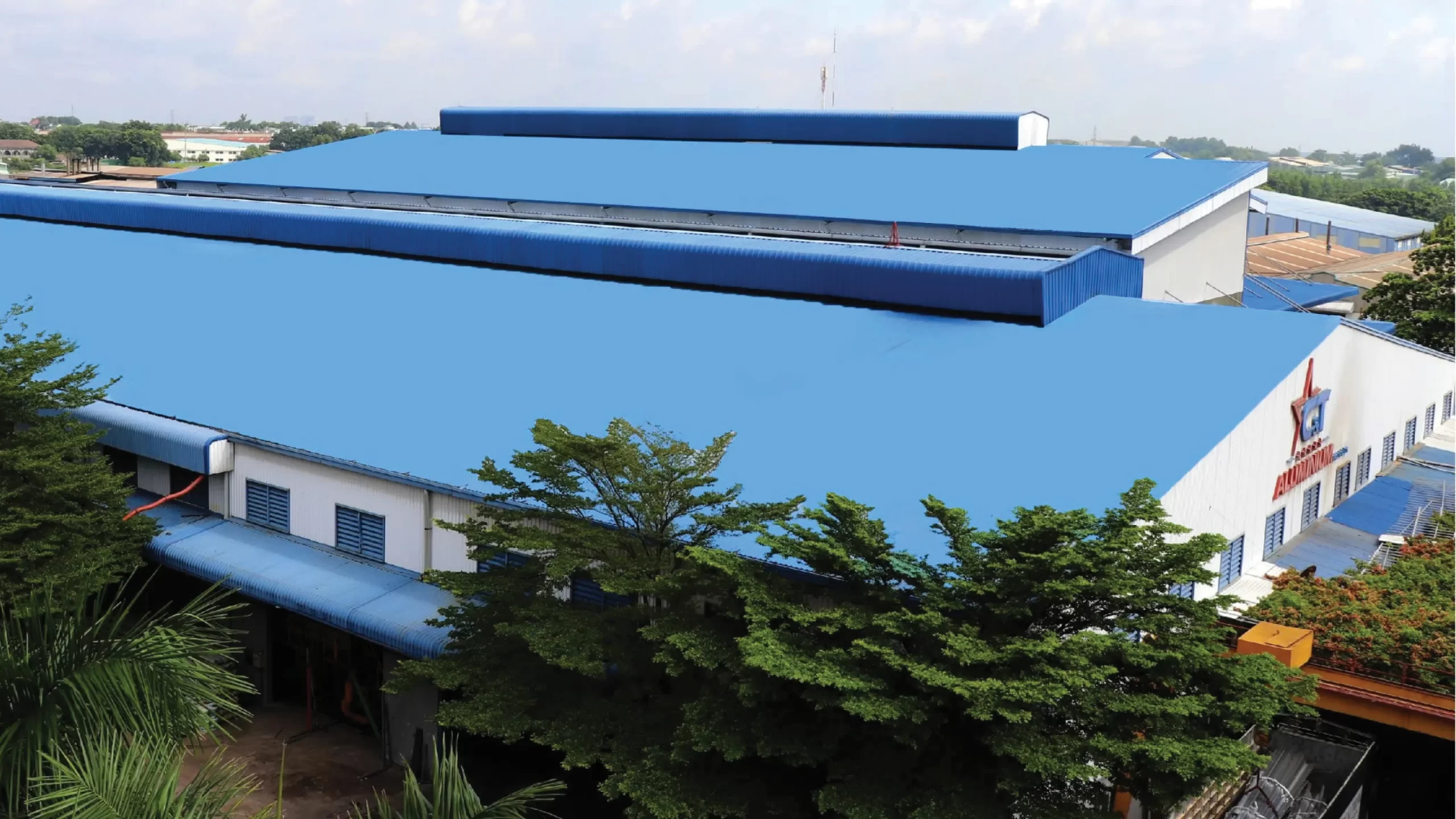 Nhà xưởng chính của Nhà máy Nhôm Cầu Thịnh với hệ thống mái xanh dương lớn, mặt trước có logo C&T Aluminium Profiles, bao quanh là nhiều cây xanh.