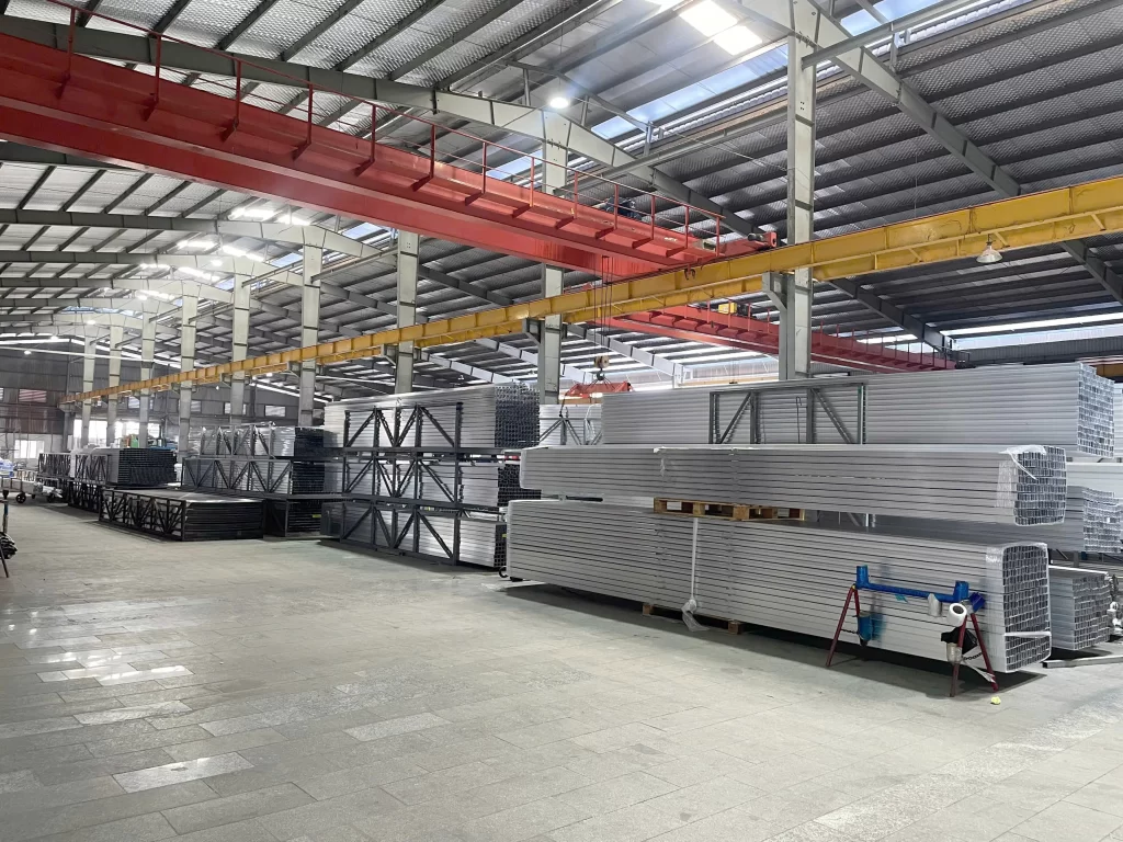 Kho nhôm công nghiệp tại nhà máy Nhôm Cầu Thịnh với các kiện thanh nhôm xếp thành chồng lớn, hệ thống cầu trục vàng đỏ phía trên.